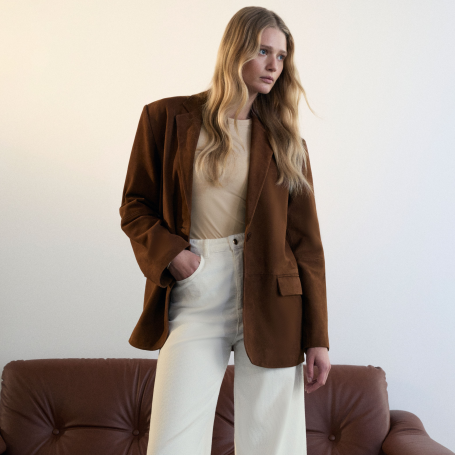 A woman wearing a a cream t-shirt, white denim jeans and a warm brown blazer.
