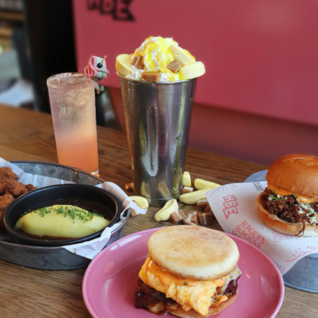 A range of yummy breakfast foods on a table. The food consists of a banana milkshake, a burger, and a breakfast muffin.