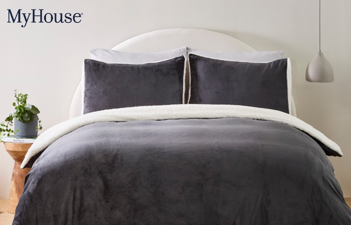 A bed with a charcoal quilt cover, matching pillows and a bedside table with a plant pot on it. Text 'MyHouse'